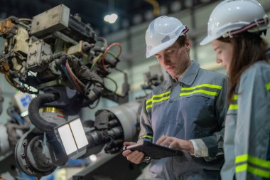 Fabrika mühendisi kadın akıllı tabletle makineyi inceliyor. İşçi robot kolunda çalışıyor. Endüstriyel bir fabrikada uzaktan kumandalı kaynak makinesi. Yapay zeka kavramı.