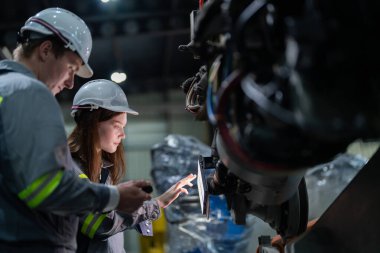 Fabrika mühendisi kadın akıllı tabletle makineyi inceliyor. İşçi robot kolunda çalışıyor. Endüstriyel bir fabrikada uzaktan kumandalı kaynak makinesi. Yapay zeka kavramı.