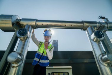 Şehir çatısı, genç bir mühendisin radyo ile güneşli bir şehir ortamında ekipmanları denetlediğini ve ayarladığını gösteriyor. İş yerinde güvenlik ve hassasiyet.