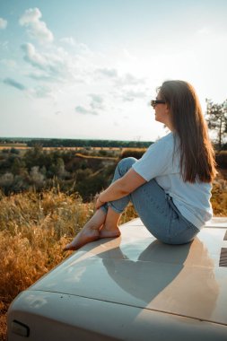 Gün batımında eski Lada arabasıyla doğada poz veren köylü kızı.