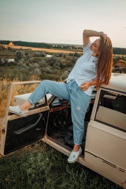 Gün batımında eski Lada arabasıyla doğada poz veren köylü kızı.