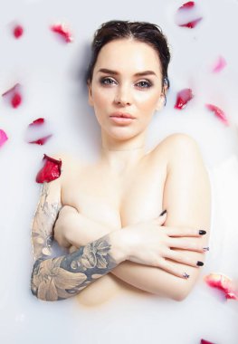 Dövmeli esmer, süt ve gül yapraklarıyla dolu bir küvette yatıyor. Göğüslerini elleriyle kapatıyor.