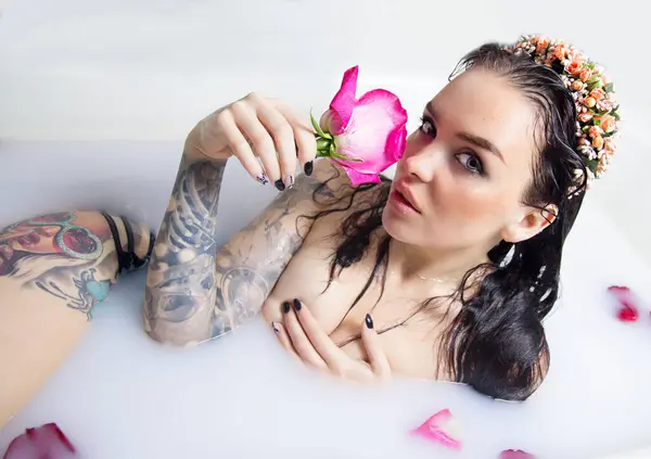a girl with a tattoo in lingerie lies in a milk bath with rose petals and holds a rose near her face