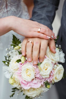 Yeni evlilerin elinde alyanslar. Gelin ve damat bir buket çiçeğin üstünde el ele tutuşuyorlar. Düğün buketi yakın plan.