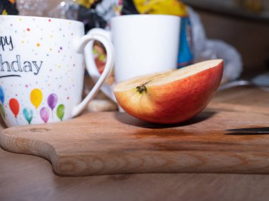 Masanın üzerinde beyaz bir fincanın yanında bıçakla kesilmiş olgun bir elma. Elmanın yarısı mutfak masasında. Bardaklar.