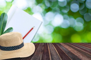Kalem ve hasır şapkalı defter yeşil bokeh arkaplanlı ahşap masada.