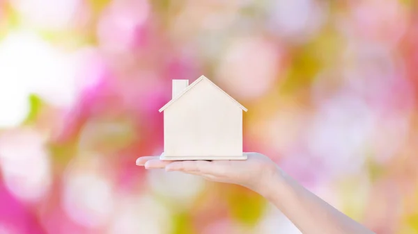 Woman hand holding small home model wooden with pink bokeh abstract background. Family life and Business real estate concept,Eco house