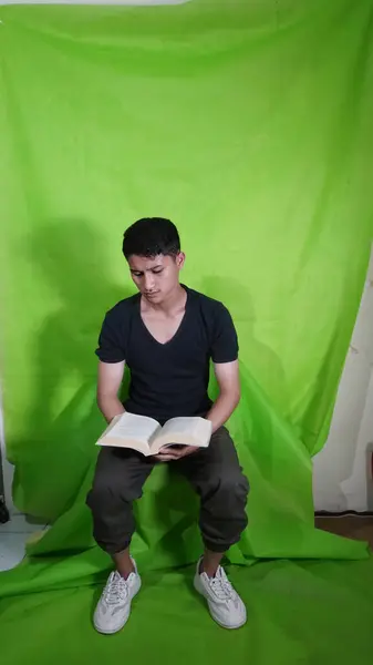 ハンサムなアジアの若者は 背景に緑色のピカンで携帯電話を再生しながら本を開いている間に座っています — ストック写真
