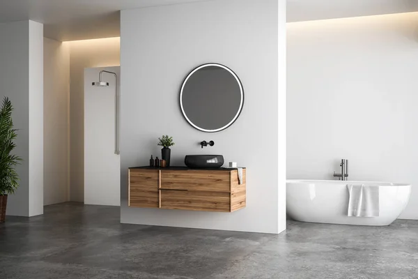 Bright bathroom interior with white tones wall, concrete floor, wooden vanity with black sink and oval mirror, white bathtub, panoramic windows.