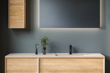 Oval lavabosu olan şık bir banyo, boş tezgah, ahşap kibir, siyah çerçeveli ayna, çiçek ve mavi duvar. Ürünlerinizi şık ve modern bir ortamda sergilemek için ideal..