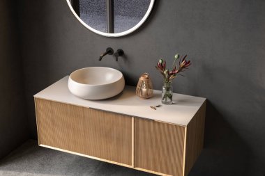 Oval lavabosu olan şık bir banyo, boş tezgah, ahşap kibir, siyah çerçeveli ayna, çiçek ve siyah duvarlar. Ürünlerinizi şık ve modern bir ortamda sergilemek için ideal..