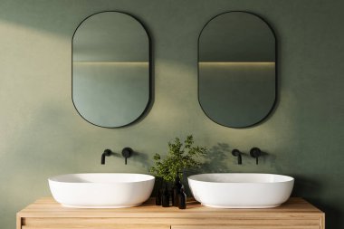 Duble oval aynaları, beyaz lavaboları ve siyah muslukları olan, sessiz yeşil bir duvara ve ahşap tezgaha dayanan gösterişli ve modern bir banyo süsü. Sakin, üç boyutlu bir canlandırma için yeşillik geliştirilmiş.