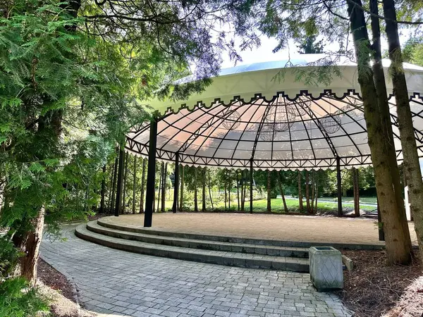 A large wrought iron dome gazebo with a canopy top with a stage in the park in the middle of green trees for events and concerts on a sunny day.