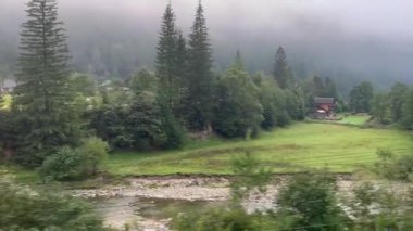 Sabah sisinin içinden geçen bir trenin yolcu vagonunun açık penceresinden turistik otel evleri ve kayalık bir kıyı boyunca akan bir nehirle birlikte dağ manzaralı vadisinin manzarasına bakın. Dinlen.