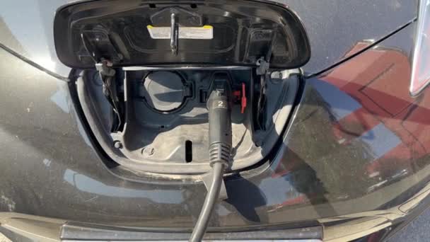 在阳光明媚的一天 一辆黑色电动车在一个充电站充电 车上车盖中央的充电器插上了电缆 电动汽车的电动汽车充电站 — 图库视频影像