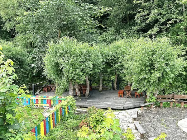 Relaxation area on a wooden terrace with dining tables and wooden chairs with a grill among green trees in the middle of the forest in summer, garden party. A view of a cozy summer garden with tables for picnics and romantic dinners.