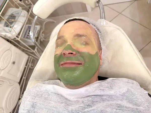 Smiling face of young man with closed eyes covered with rejuvenating green mask in spa salon. A modeling lifting mask is applied to the entire face of a man lying on a couch in a cosmetology clinic.