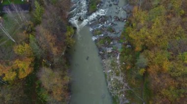 İnsansız hava aracı sonbahar ormanının, nehrin ve evin üzerinde ağaçların arasında uçuyor. Ağaçlarda renkli yapraklar olan orman ve dağ nehri manzarası, üst manzara. Geniş bir nehrin arka planındaki kayalık akıntılı sonbahar ağaçlarının en üst görüntüsü.