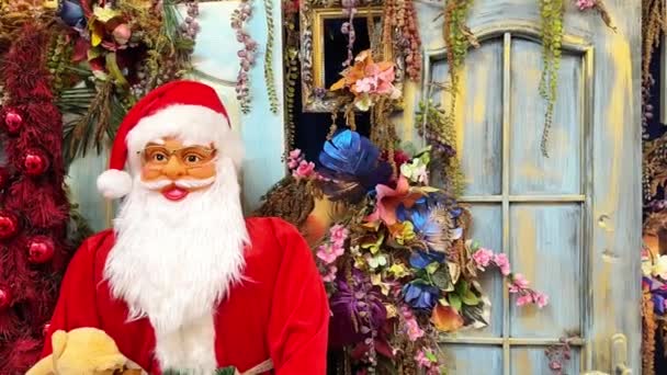 圣诞老人手里拿着一个柔软的玩具 站在商店门口 装饰着圣诞装饰品 迎接客人 展品上有个有趣的玩具圣诞老人 特写镜头 — 图库视频影像