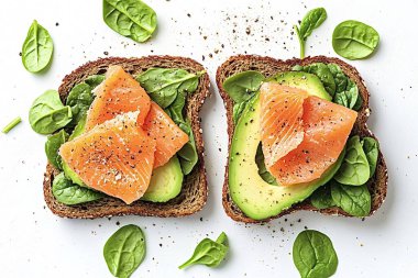 İki dilim tam tahıllı tost avokado ve füme somon. Karabiberle tatlandırılmış ve taze ıspanak yapraklarıyla süslenmiş beyaz arka planda yatıyor.