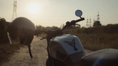 Motosikletin gidonuna takılı gümüş bir kask sürücüyü bekliyor. Gün batımı zamanı