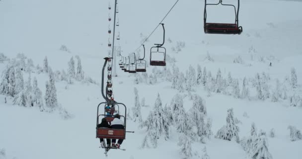 チェアリフトはスキーヤーとスノーボーダーを曇った日に山の頂上に連れて行きます 雪に覆われた山の風景 — ストック動画
