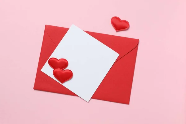 Red paper envelope with blank white note mockup inside and red hearts on pink background. Flat lay, top view
