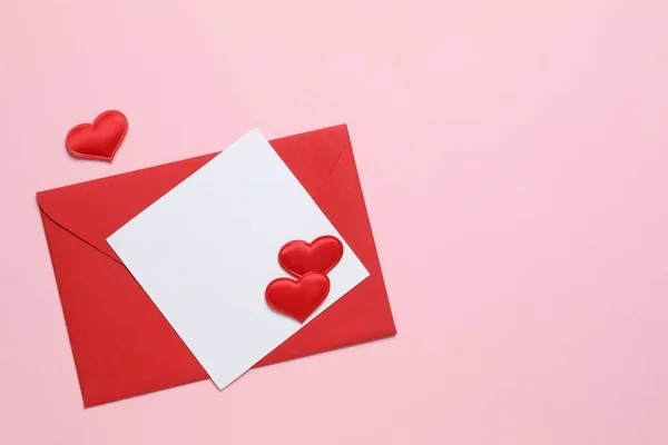 Red paper envelope with blank white note mockup inside and red hearts on pink background. Flat lay, top view