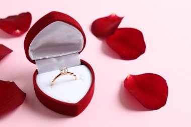 Güzel, parlak, altın bir nişan yüzüğü. Büyük elmas değerli taşıyla birlikte. Arka planda gül yaprakları olan kırmızı bir kutuda.