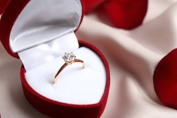 美丽的金光闪闪的订婚戒指 镶嵌着巨大的钻石宝石 镶嵌在缀有玫瑰花瓣的丝织品上华丽的红色天鹅绒盒中 图库照片