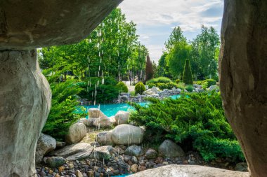 Stone ve Falling Water 'ın Çerçevelettiği Bahçe Sahnesi. Taş bir şelalenin arkasından manzaralı güzel bir bahçe vahası. Kayalık bir çıkıntı. Su, el değmemiş mavi bir gölete akıyor. Seçici bir odak noktası.