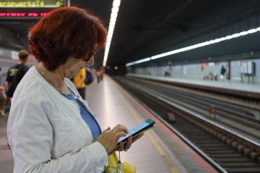 Yaşlı kadın, şehir içinde tren beklerken kırsal kesime gidip telefona bakıyor ve bekliyor.