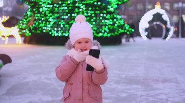 Küçük kız sokakta Noel ışıkları ağacının yanında duruyor, akıllı telefondan konuşuyor ve tebrik ediyor. İnternetten tebrik ederim.