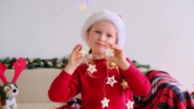 Noel Baba şapkalı gülümseyen kız kameraya bakıp Noel ışıklarıyla komik surat yapıyor. Noel kızının portresi.