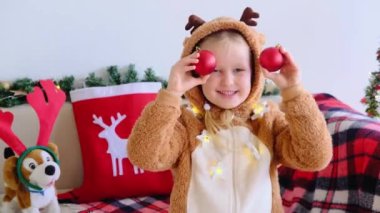 Geyik kostümlü gülümseyen çocuk kameraya bakıp yüzünde kırmızı Noel toplarıyla komik bir ifade takınıyor. Noel kızının portresi.