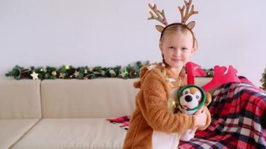 Geyik kostümlü gülümseyen çocuk ve köpek kameraya bakıp dans ediyorlar. Noel kızının portresi