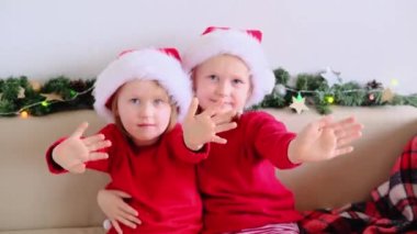 İki mutlu kız kardeş Noel Baba şapkalı çocuklar el sallıyor ve kameraya bakıyor. 4k Christmas online Evden tebrikler.
