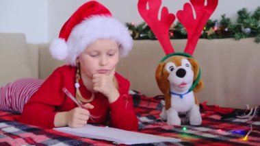 Noel Baba şapkalı mutlu küçük kız evde yatakta yatıyor ve Noel Baba 'ya hediyeler için mektuplar yazıyor. Noel mucizesi dilek listesi. Noel ışıkları olan çocuk