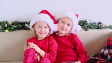 İki mutlu kız kardeş Noel Baba şapkalı çocuklar el sallıyor ve kameraya bakıyor. 4k Christmas online Evden tebrikler.