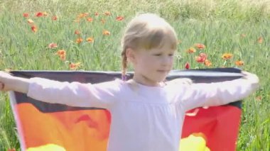 A little girl with a Germany flag on her back walks on a wheat field with poppies in summer. Flag of Germany.