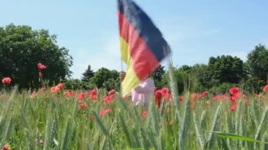 A little blond girl with a Germany flag on her hands on a green wheat field with poppies in summer. Flag of Germany. patriotism, independence day