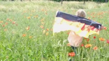 A little girl with a German flag on her back walks on a wheat field with poppies in summer. Flag of Germany.