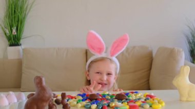 Little blond girl with rabbit ears hunt for chocolate eggs eat Kid celebrating Easter. Child on Easter egg hunt.