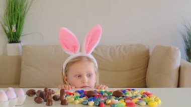 Little blond girl in bunny ears find and pick up multicolored egg on Easter egg hunt. Looks surprised at Easter chocolate eggs.
