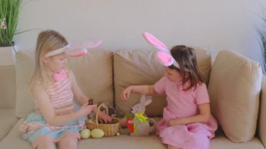 Two little girls sisters in rabbit ears sit on the couch and share found chocolate eggs with each other. Easter egg hunt. Happy easter childhood