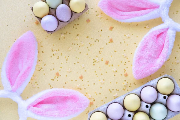 Colorful Easter eggs with rabbit ears and confetti on yellow background. Egg hunting. Flat lay, top view, copy space.