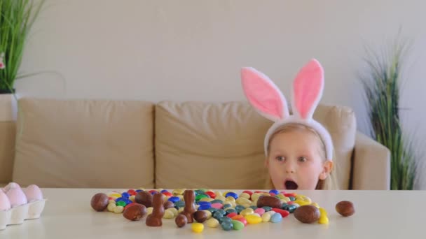 ウサギの耳の中の小さな金髪の女の子は幸せそうで テーブルの上にチョコレートの卵をたくさん抱いています イースターエッグハント 幸せなイースターの子供時代 — ストック動画