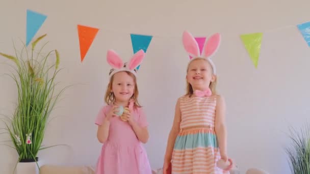 两个快乐的小女孩在兔子耳朵里跳着舞 抱着复活节彩蛋在沙发上跳舞 有趣的疯狂快乐的宝贝 复活节给孩子 — 图库视频影像