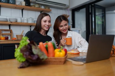 Beautiful woman and friend talking and using laptop at kitchen room. Girl friends couple living together at home