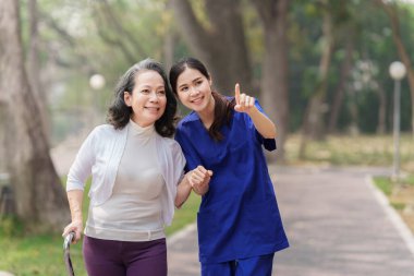 Sağlık hemşiresi, dışarıda yaşlı bir kadınla fizik tedavi. Hemşire el ele tutuşup yaşlı kadının yürümesine yardım ediyor..